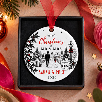 A round white Christmas ornament with a red ribbon hanger, displayed inside a black gift box. The ornament features a black-and-white illustration of a bride and groom walking under a tree, with red text reading 'Our Finest Christmas as Mr & Mrs Sarah & Mike 2026'.