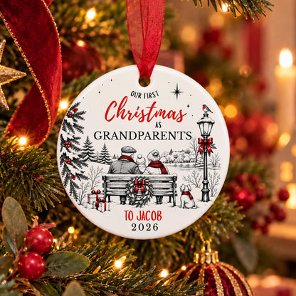 A personalized round ceramic Christmas ornament hanging on a decorated Christmas tree. The white ornament features a black and red illustration of a grandfather and grandmother sitting on a bench under a streetlamp, with the text 'OUR FIRST Christmas AS GRANDPARENTS TO JACOB 2026'.