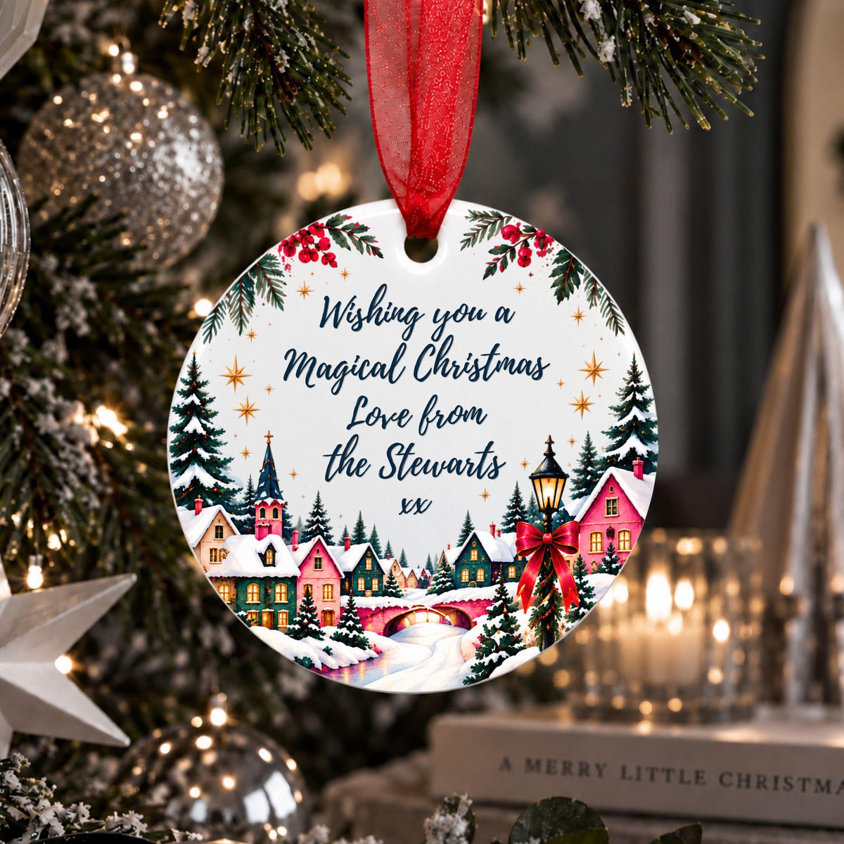 A round Christmas ornament hanging from a red ribbon on a decorated tree. The ornament features a winter village scene with colorful houses, evergreen trees, stars, and the text, 'Wishing you a Magical Christmas Love from the Stewarts xx'.