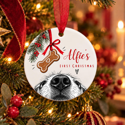 A round ceramic Christmas ornament hanging on a decorated Christmas tree. The ornament is white with a red ribbon, featuring a black-and-white dog face illustration, a red ribbon bow, a brown bone-shaped cookie graphic with the word 'Love', and the text 'Alfie's First Christmas'.