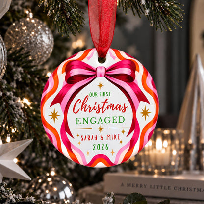 A personalized round Christmas ornament hanging from a red ribbon in a decorated Christmas tree. The ornament features a colorful wavy pattern in pink, orange, white, and red, with a pink bow illustration at the top and custom text reading 'OUR FIRST Christmas ENGAGED Sarah & Mike 2026'.